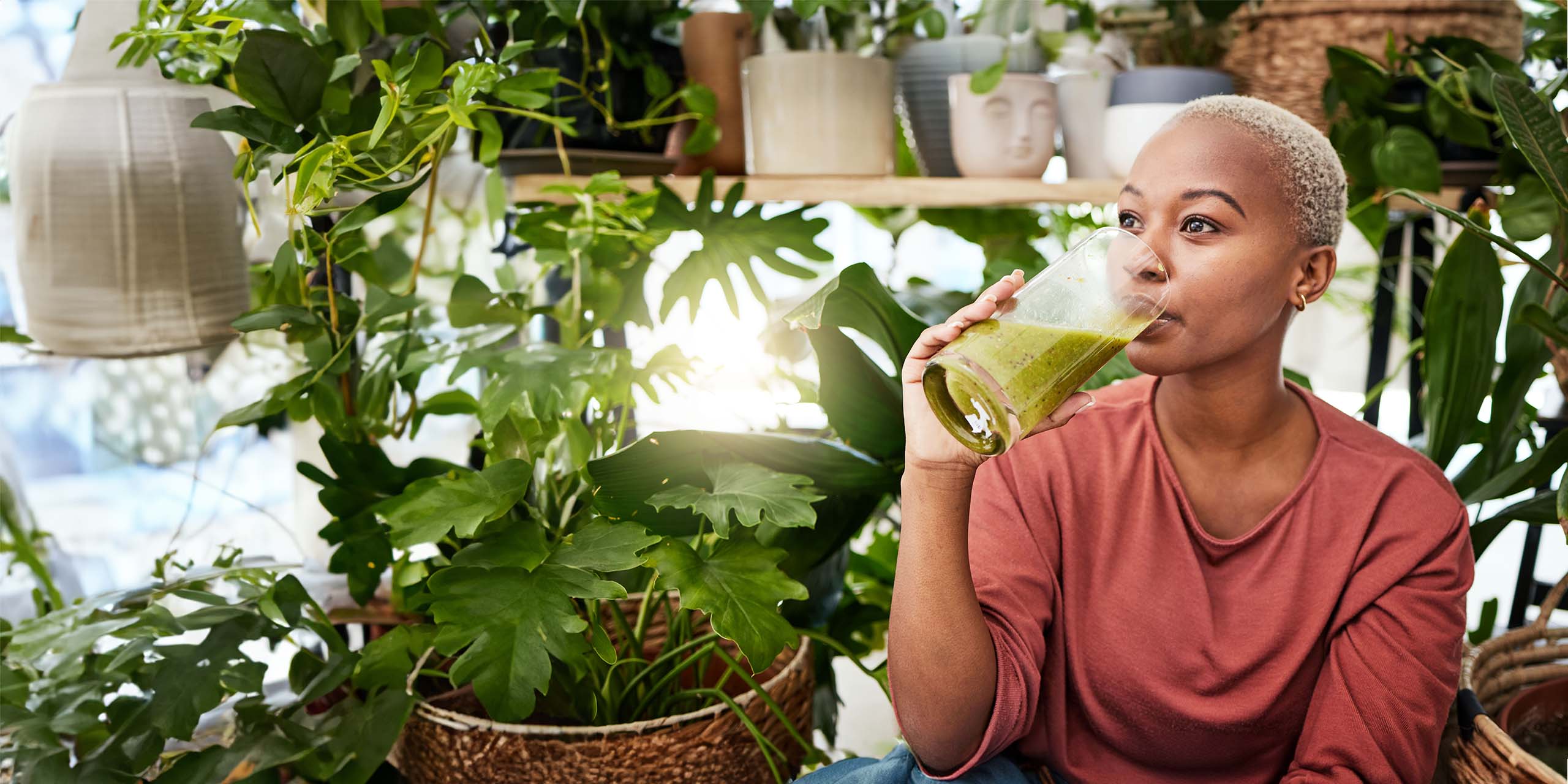 Ein Frau umgeben von Zimmerpflanzen trinkt einen grünes Smoothie.