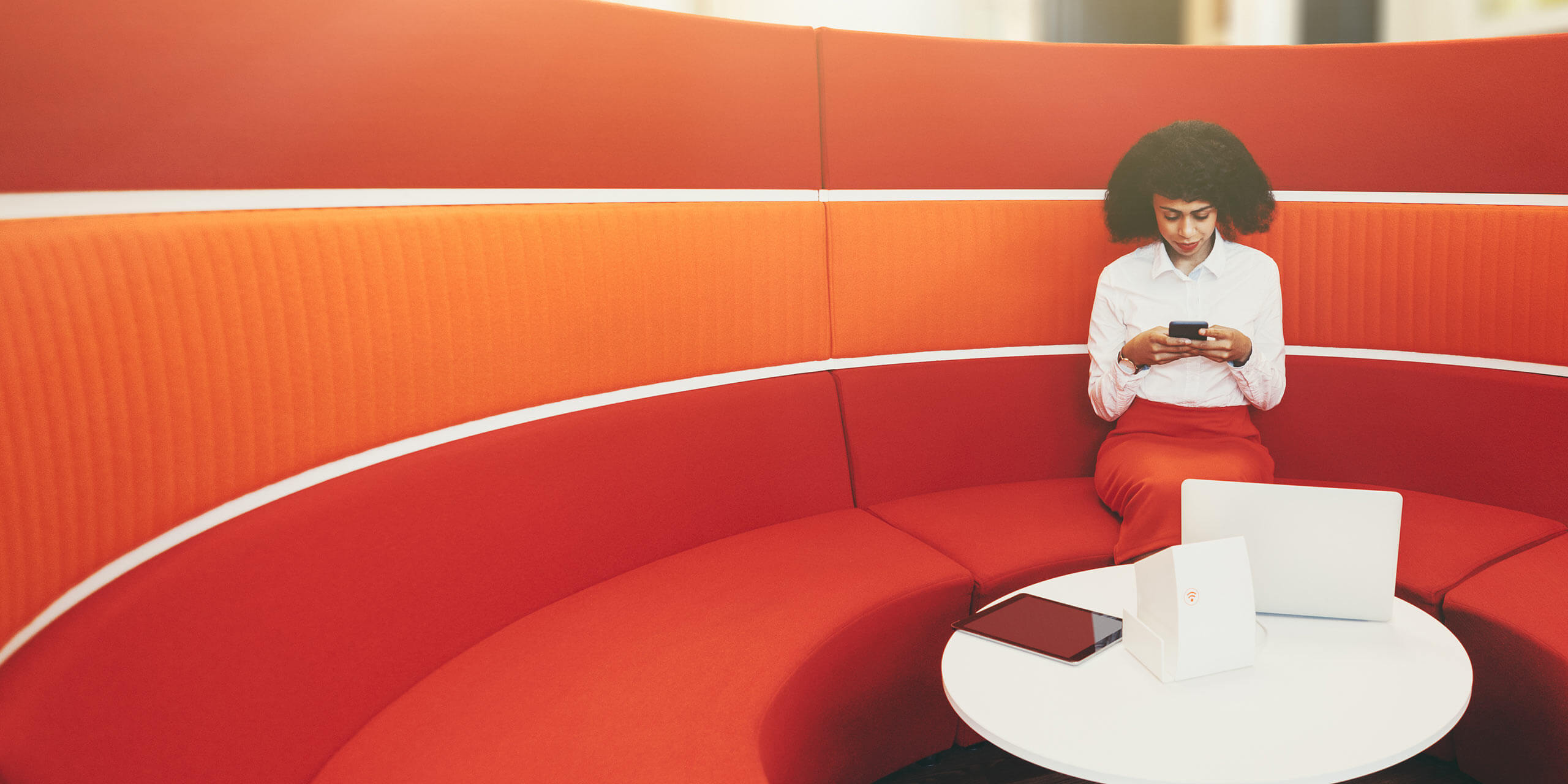 Eine Person mit lockigem Haar, bekleidet mit einem weißen Hemd und einem roten Rock, sitzt auf einem orangefarbenen, U-förmigen Sofa und benutzt ein Smartphone. Auf einem weißen Tisch vor ihr liegen ein Laptop, ein Tablet, ein Ordner und Papiere. Der Hintergrund zeigt ein minimalistisches, modernes Interieur.