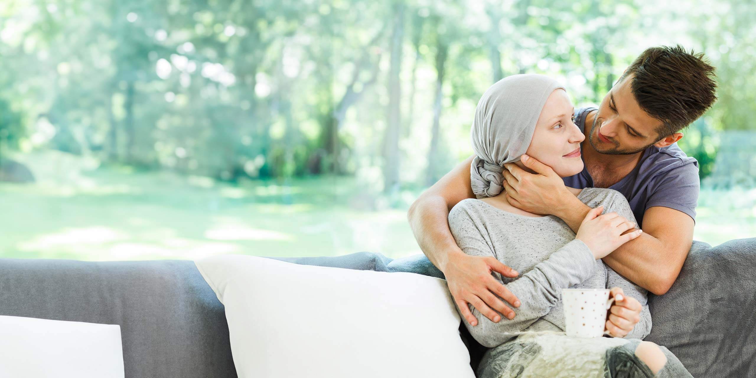 Ein Mann umarmt zärtlich eine Frau mit Kopftuch, die auf einem Sofa in einem ruhigen, sonnenbeschienenen Garten sitzt. Die Frau hält mit beiden Händen einen weißen Becher und beide blicken sich nachdenklich und liebevoll in die Augen.